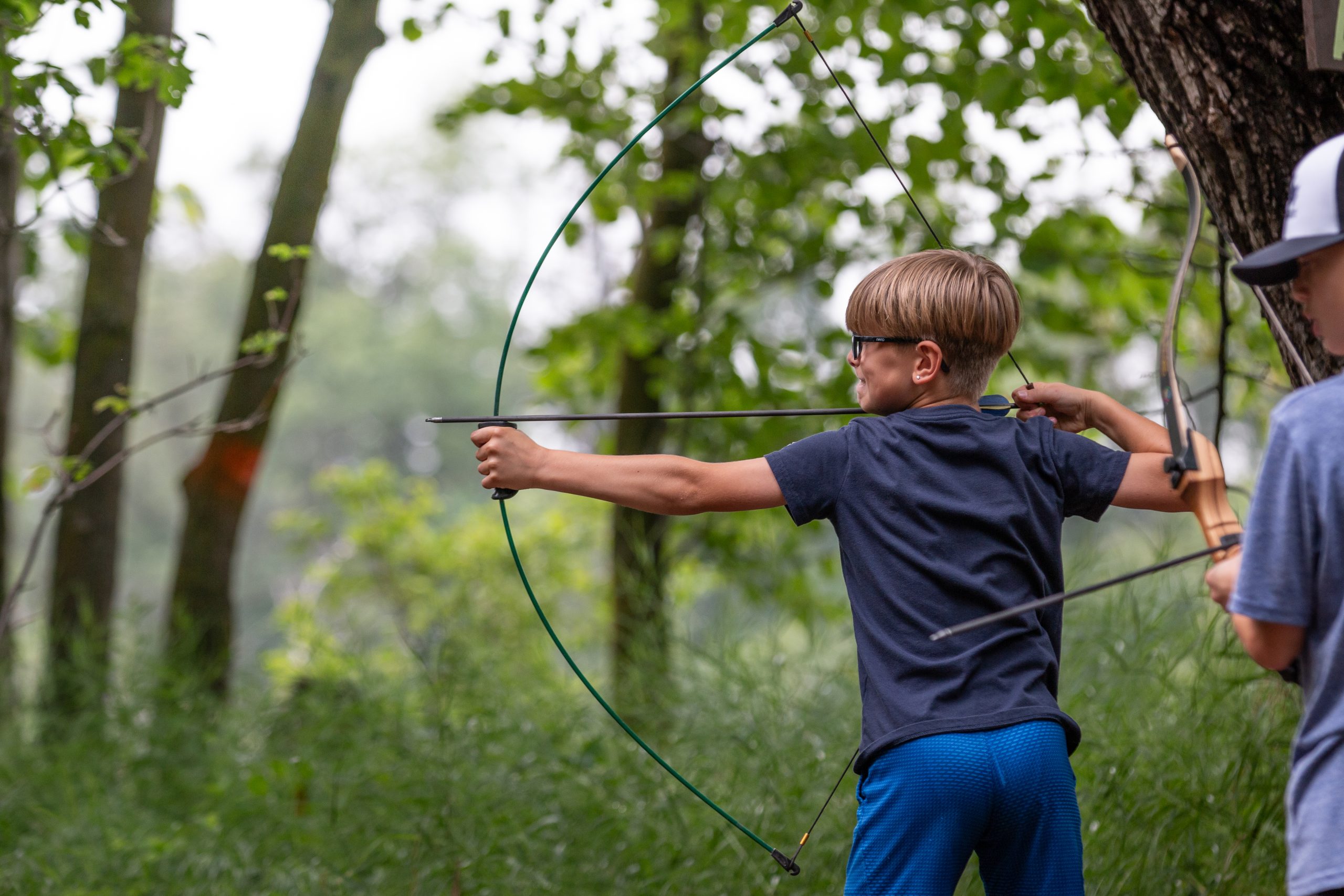 Boy in an area with green trees, pulling back a bow with an arrow in it.